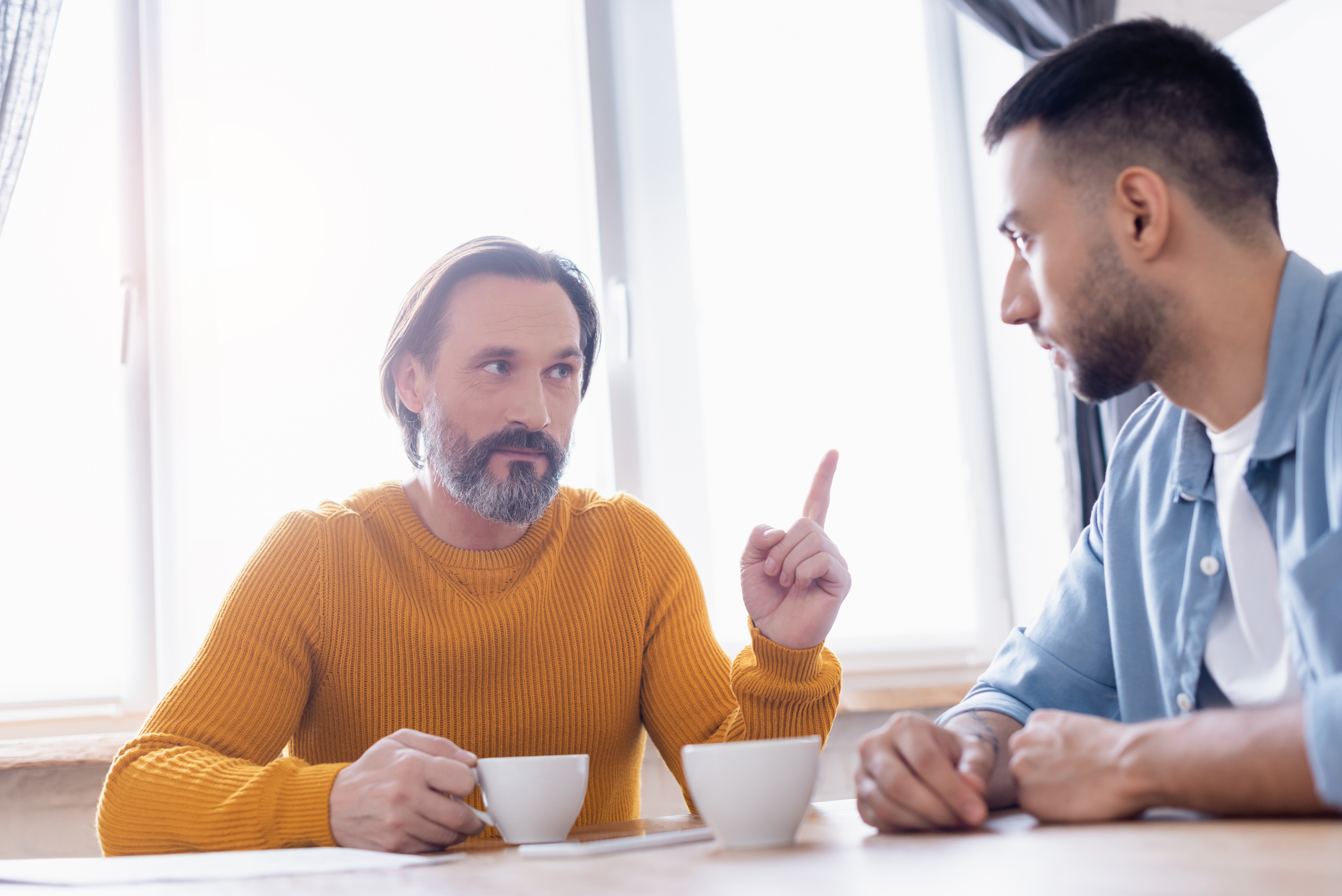 Two men sitting at a table with coffee cups, engaged in conversation. The man on the left, wearing a mustard sweater, raises his index finger, seemingly making a point. Sunlight streams through a window behind them.