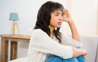 A woman sits on a couch, resting her chin on her hand with a thoughtful expression. She is in a bright room with soft lighting, and a wooden table with a small lamp is visible in the background.