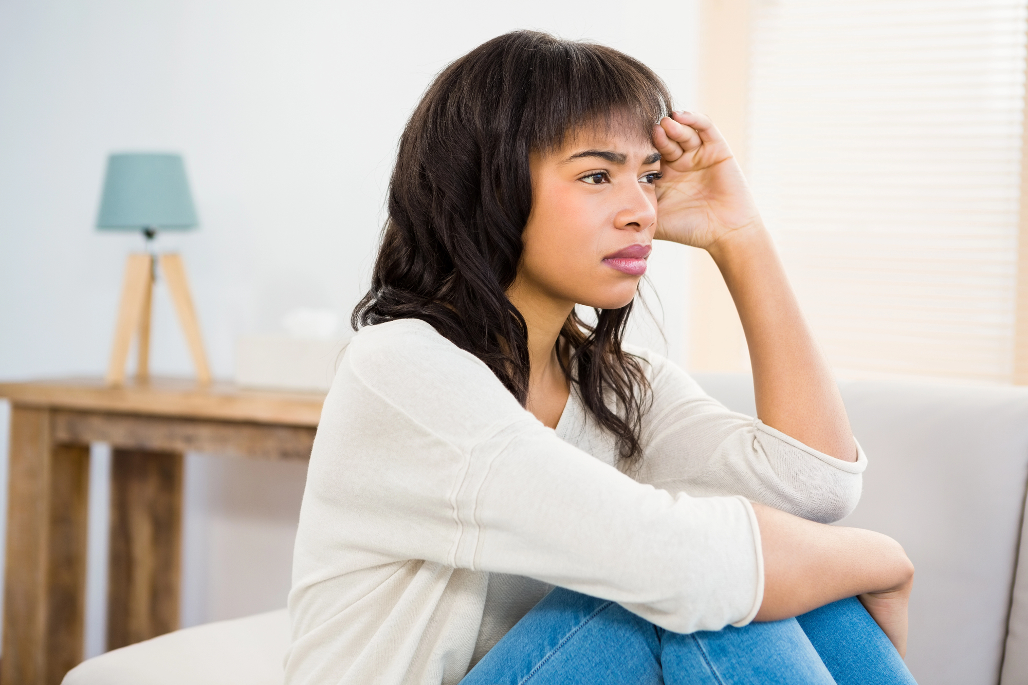 A woman sits on a couch, resting her chin on her hand with a thoughtful expression. She is in a bright room with soft lighting, and a wooden table with a small lamp is visible in the background.