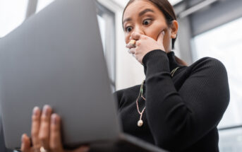 A surprised woman with long hair and a black turtleneck holds a laptop, covering her mouth with her hand, standing in a bright room with large windows.