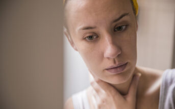 A person with a thoughtful expression looks down while touching their neck. They have light skin and are wearing a yellow headband and a white tank top. The scene is softly lit, creating a calm and introspective atmosphere.