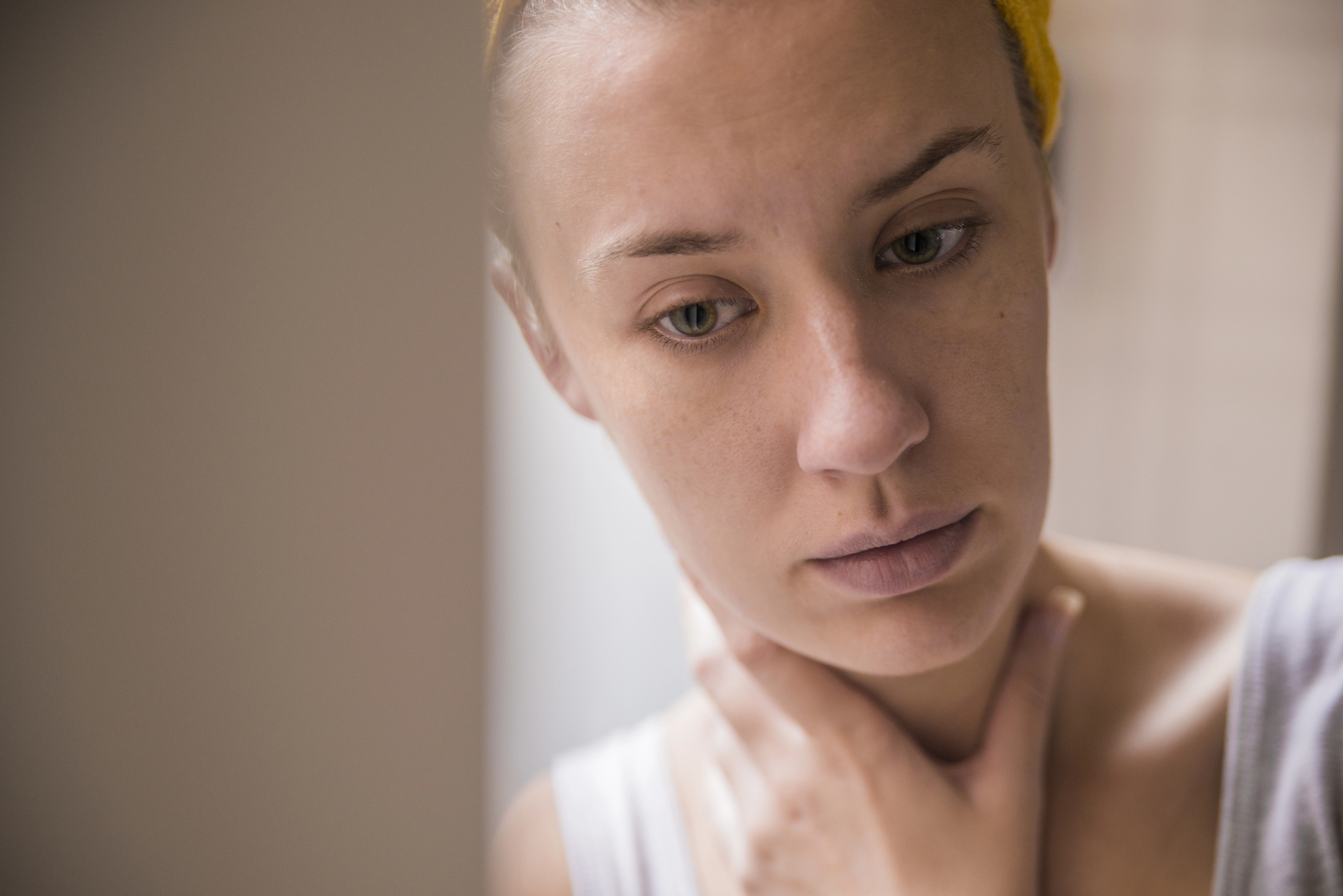 A person with a thoughtful expression looks down while touching their neck. They have light skin and are wearing a yellow headband and a white tank top. The scene is softly lit, creating a calm and introspective atmosphere.