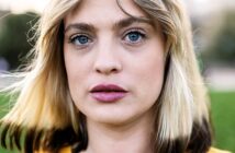 A close-up of a person with blonde hair, striking blue eyes, and wearing a yellow top. The background is blurred greenery, creating a soft focus on the subject's face and expression.