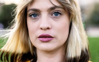 A close-up of a person with blonde hair, striking blue eyes, and wearing a yellow top. The background is blurred greenery, creating a soft focus on the subject's face and expression.