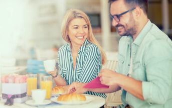 A smiling woman and man enjoy breakfast at a cafe. The woman holds a cup, while the man prepares his meal. They are seated at a table with pastries, juice, and a small gift box, surrounded by a bright and airy ambiance.