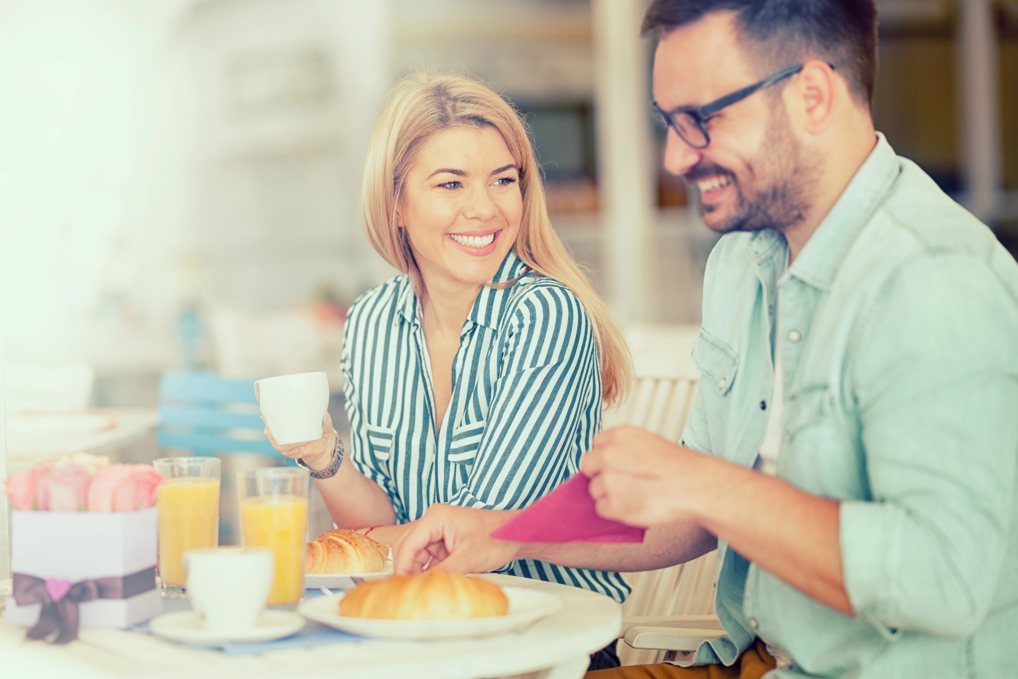 A smiling woman and man enjoy breakfast at a cafe. The woman holds a cup, while the man prepares his meal. They are seated at a table with pastries, juice, and a small gift box, surrounded by a bright and airy ambiance.