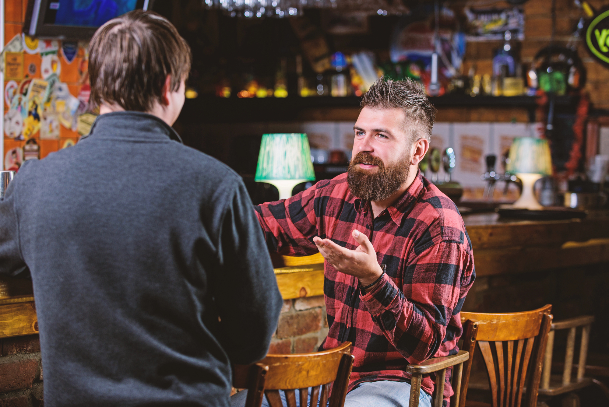 Two men sit in a bar, engaged in conversation. One has a beard and wearing a red flannel shirt, gestures with his hand while speaking. The setting features wooden chairs and a dimly lit, cozy ambiance with decorative elements.