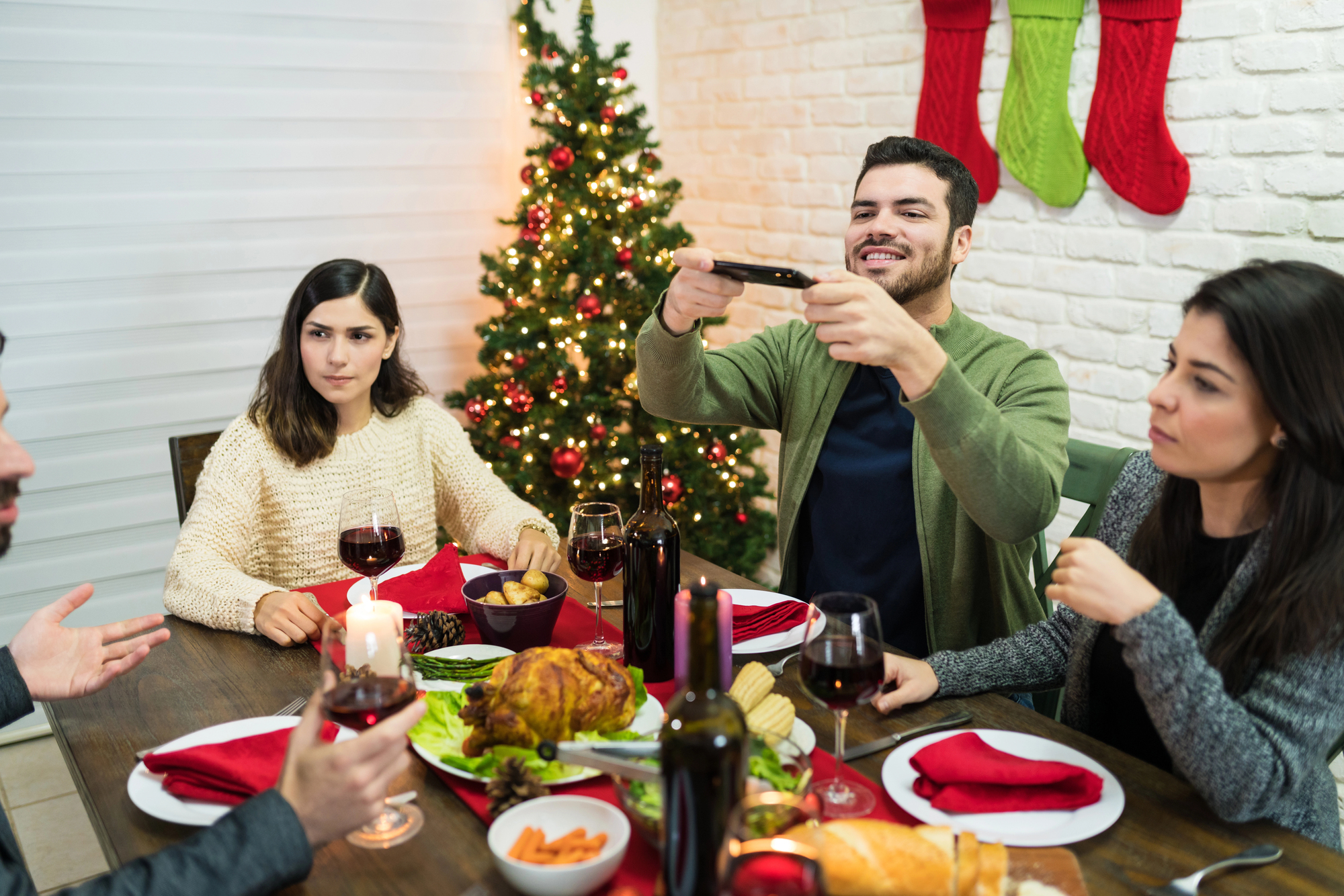 A group of four people gather around a festive dinner table with a roasted chicken, wine, and candles. A man takes a photo with his phone, while a Christmas tree and stockings decorate the background.