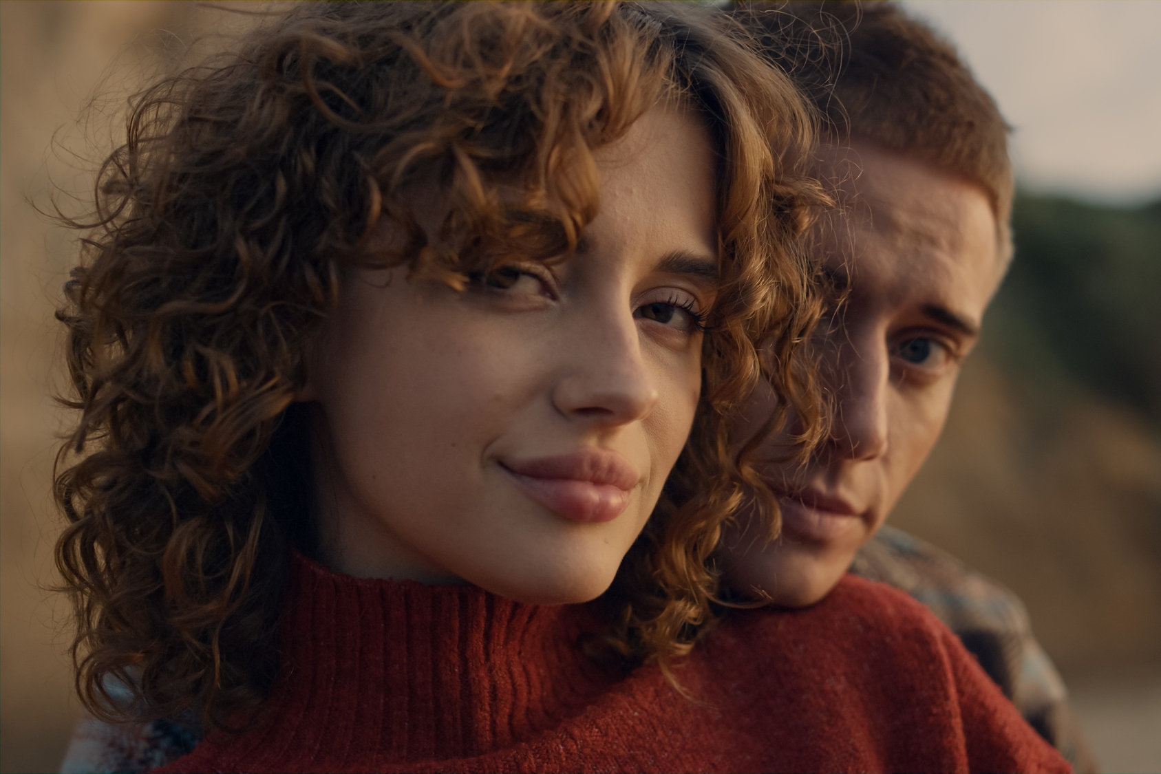 A close-up of a person with curly hair wearing a red sweater, leaning against another person with short hair. They are outdoors, with natural lighting and a blurred background. Both have neutral expressions.