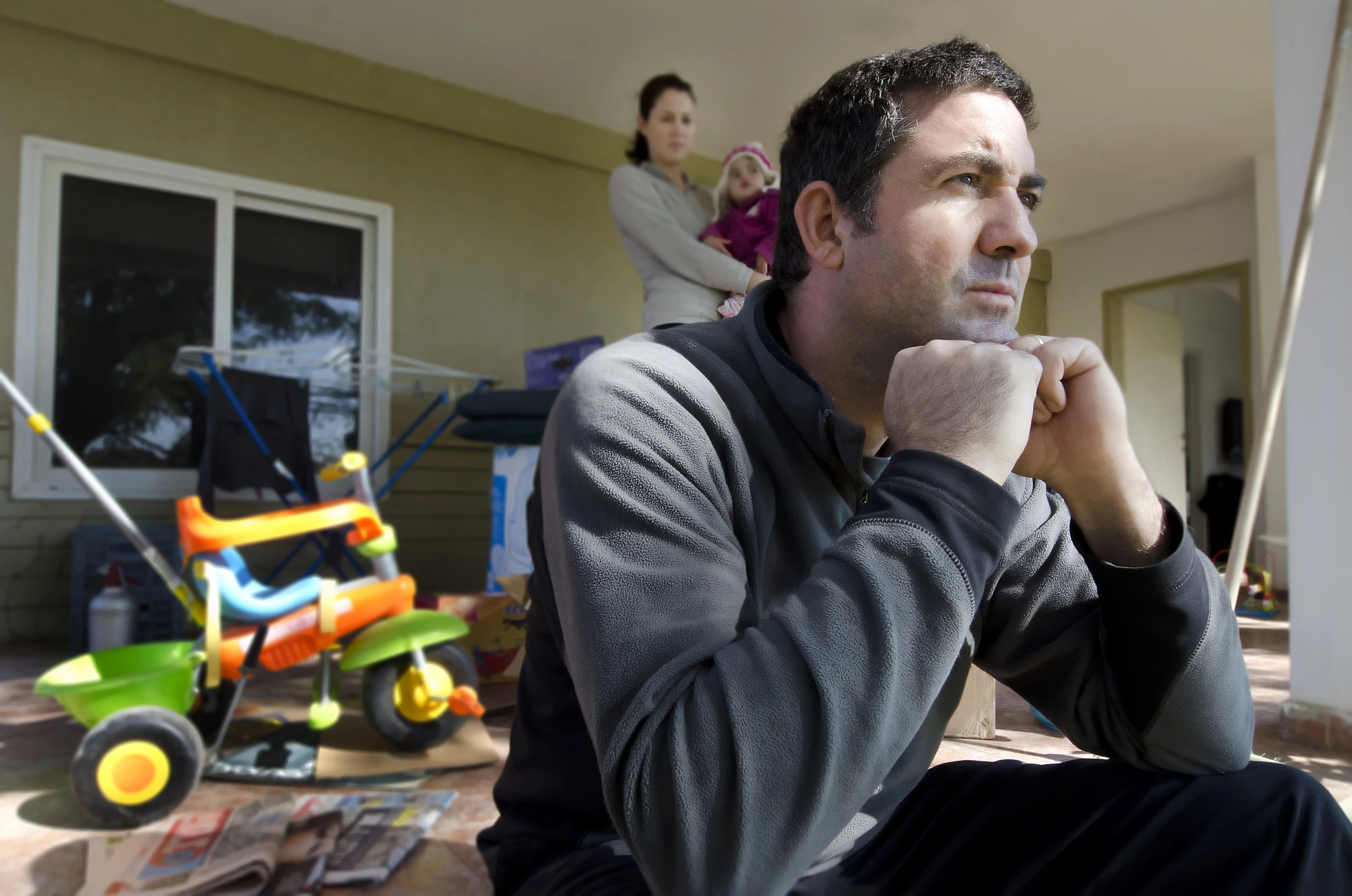 A man sits on a front porch with a thoughtful expression, resting his chin on his hands. In the background, a woman holds a child. A toy tricycle and a scattered newspaper are on the ground nearby.