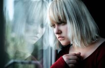 A woman with blonde hair gazes thoughtfully out a window, with her reflection visible in the glass. She rests her hand gently on the window frame, wearing a red top.