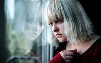 A woman with blonde hair gazes thoughtfully out a window, with her reflection visible in the glass. She rests her hand gently on the window frame, wearing a red top.