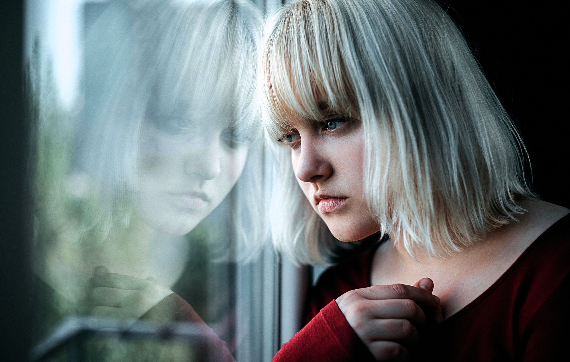 A woman with blonde hair gazes thoughtfully out a window, with her reflection visible in the glass. She rests her hand gently on the window frame, wearing a red top.