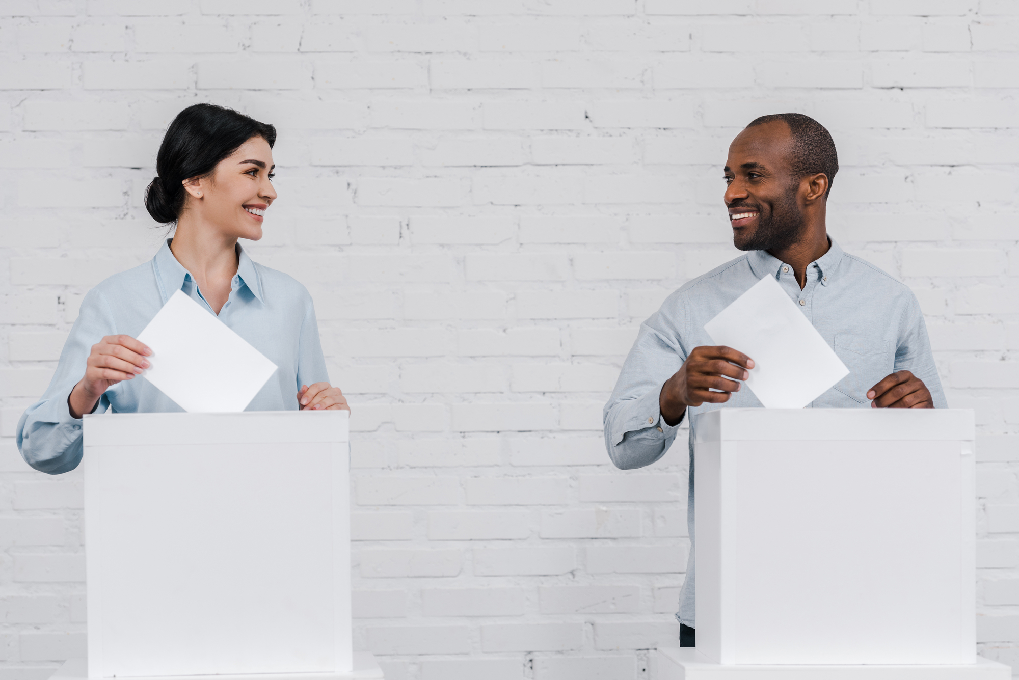 A woman and a man are smiling at each other while placing papers into separate white boxes, suggesting a voting or ballot scenario, against a white brick wall background.