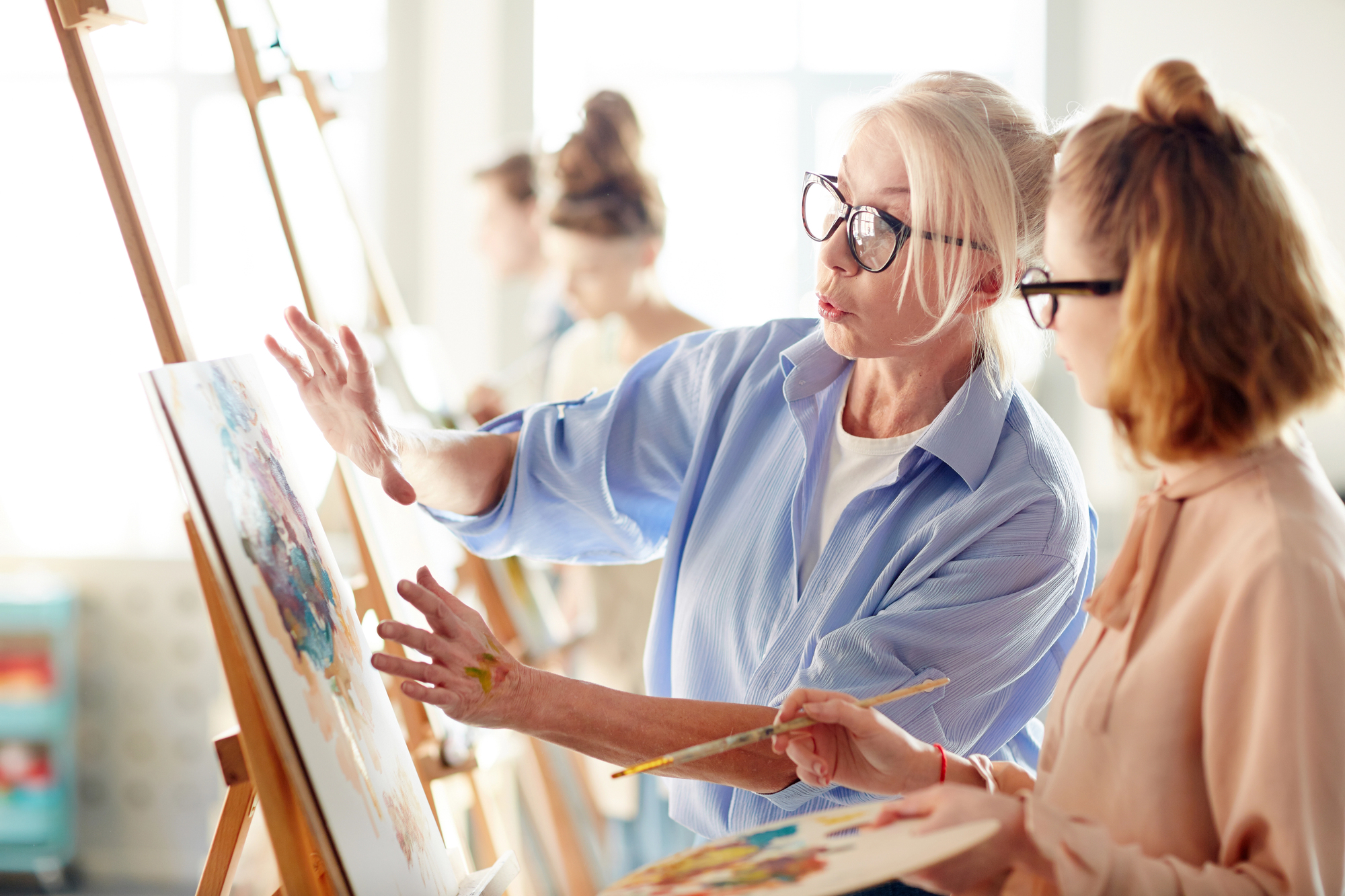 A senior woman in glasses is teaching a younger woman how to paint at an easel. The room is filled with natural light, and other students can be seen working in the background.