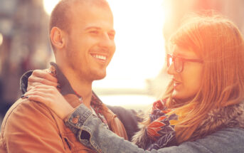 A couple stands close together outdoors, smiling at each other. The woman, wearing glasses and a scarf, has her hands on the man's shoulders. The man is wearing a brown jacket. The background is softly blurred with warm sunlight.