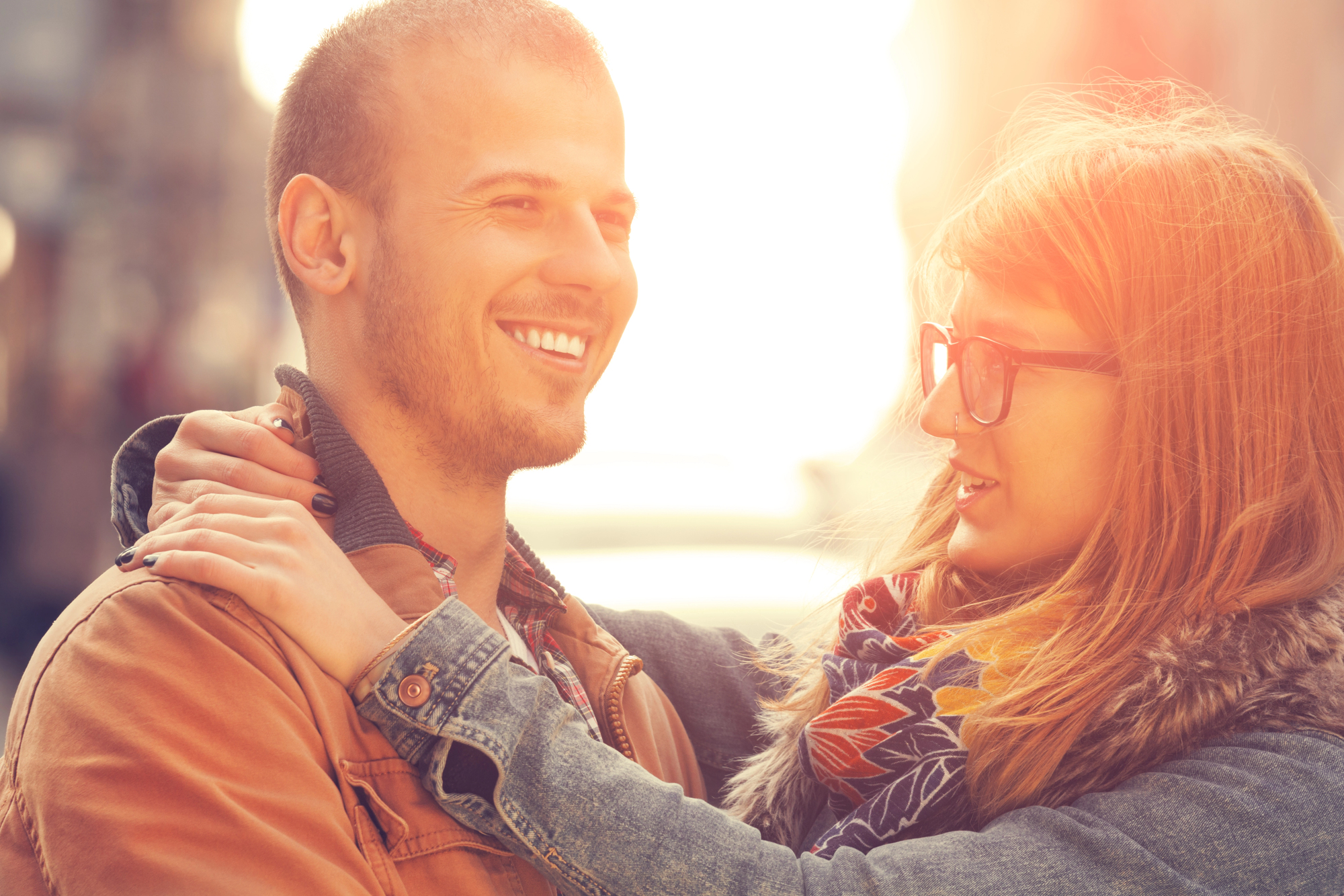 A couple stands close together outdoors, smiling at each other. The woman, wearing glasses and a scarf, has her hands on the man's shoulders. The man is wearing a brown jacket. The background is softly blurred with warm sunlight.