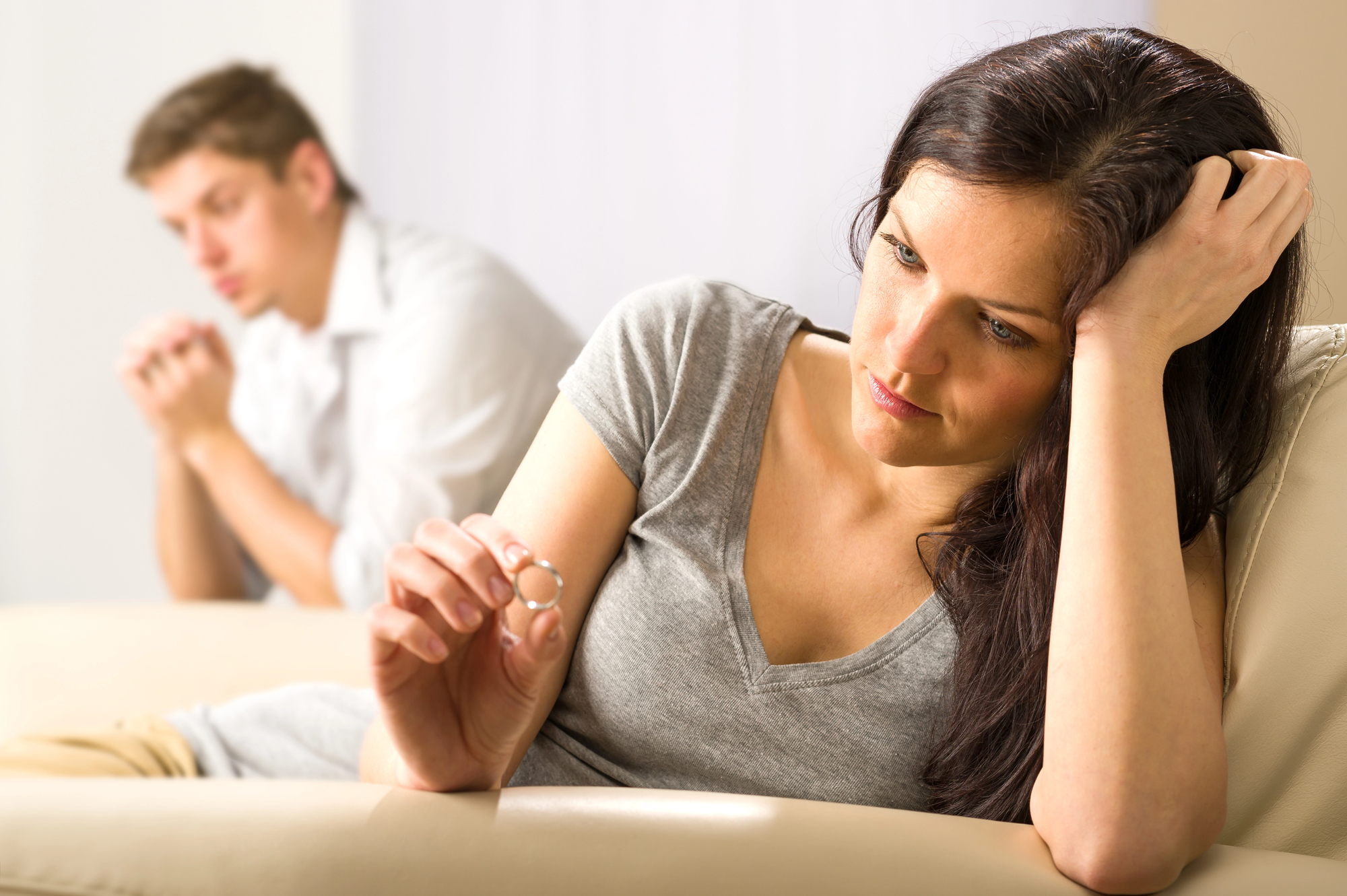 A woman sitting on a couch looks contemplatively at a ring she holds, while a man sits in the background with a thoughtful expression. Both appear pensive, creating a mood of reflection and contemplation.