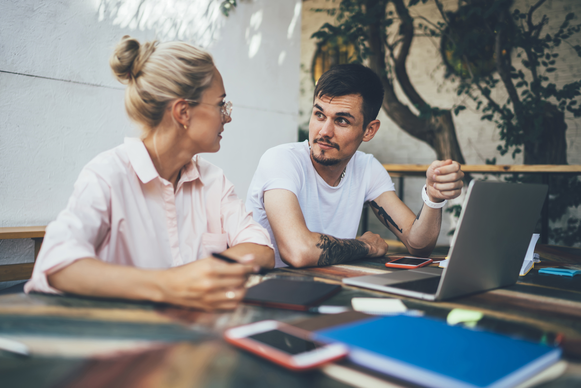 A woman and a man are sitting at an outdoor table with a laptop and notebooks. They appear to be having a discussion. The setting is casual, with trees visible in the background.