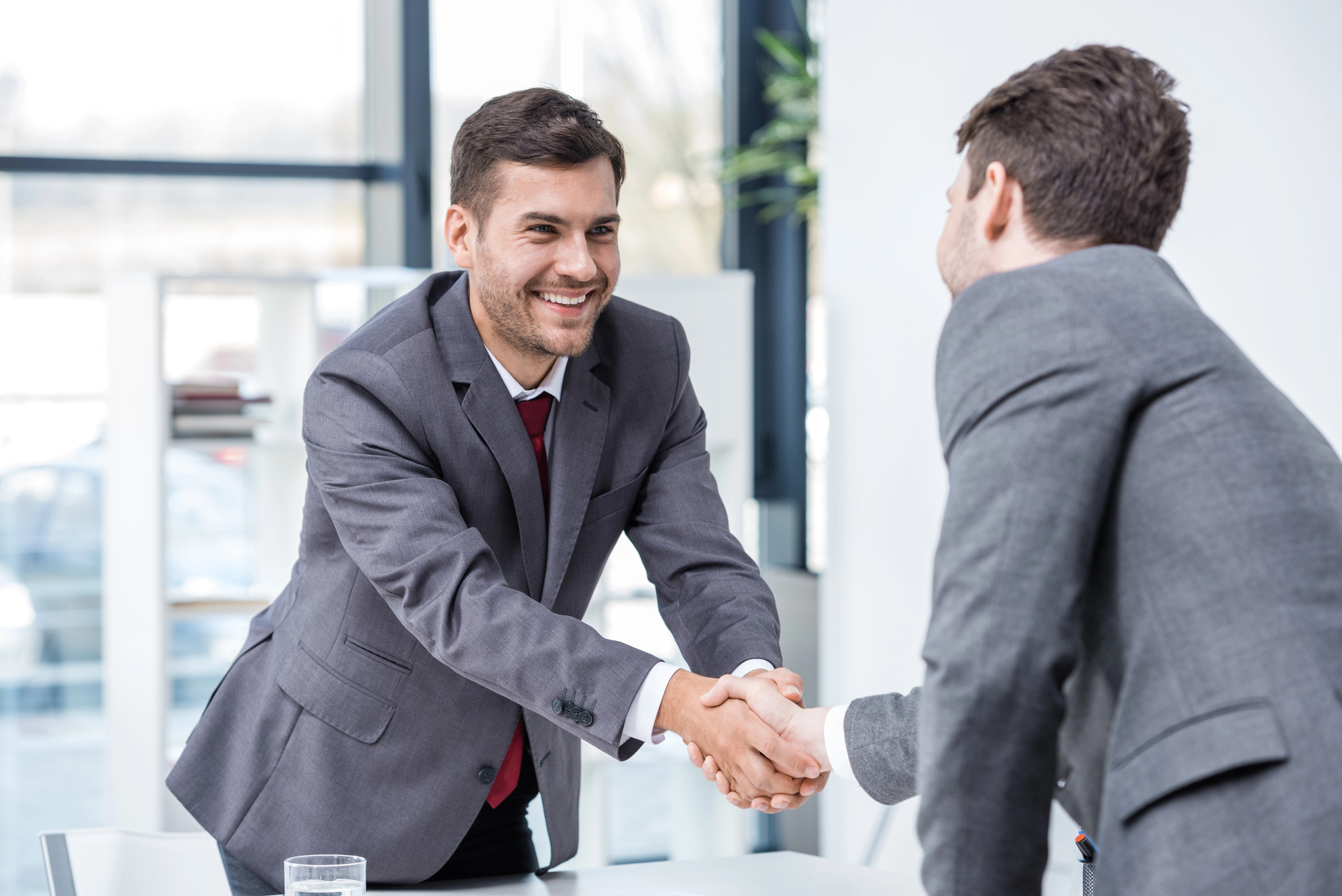 Two businessmen in suits shaking hands in a bright office setting. One man is smiling, and a table with documents and a glass of water is visible. Background shows large windows and a shelf.