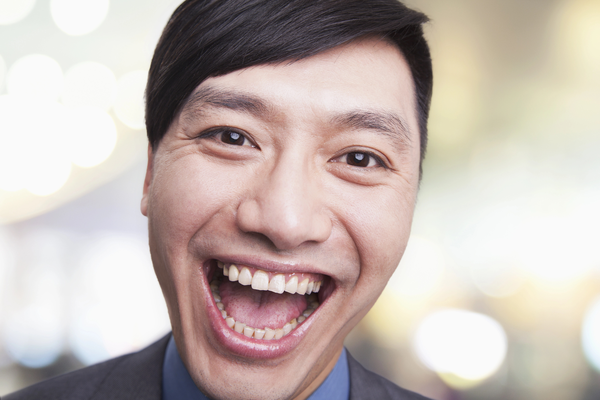 A person with short dark hair smiling widely, wearing a suit and tie. The background is blurred with bright bokeh lights, creating a cheerful and lively atmosphere.