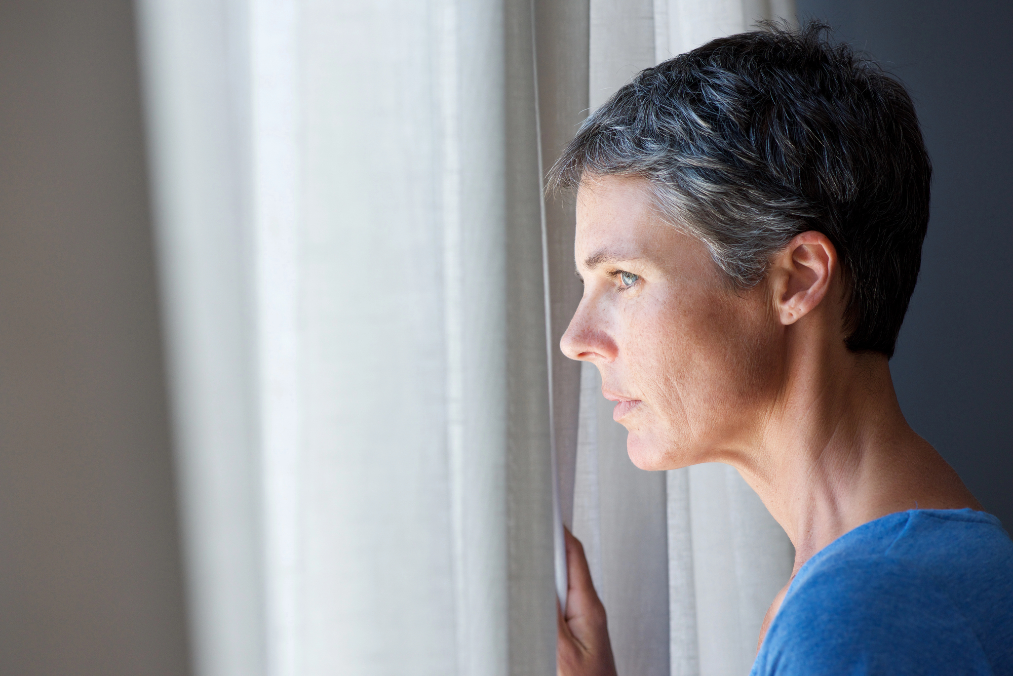 A person with short gray hair and a blue shirt looks out of a window with a thoughtful expression. They hold the white curtain gently, as soft daylight illuminates one side of their face.