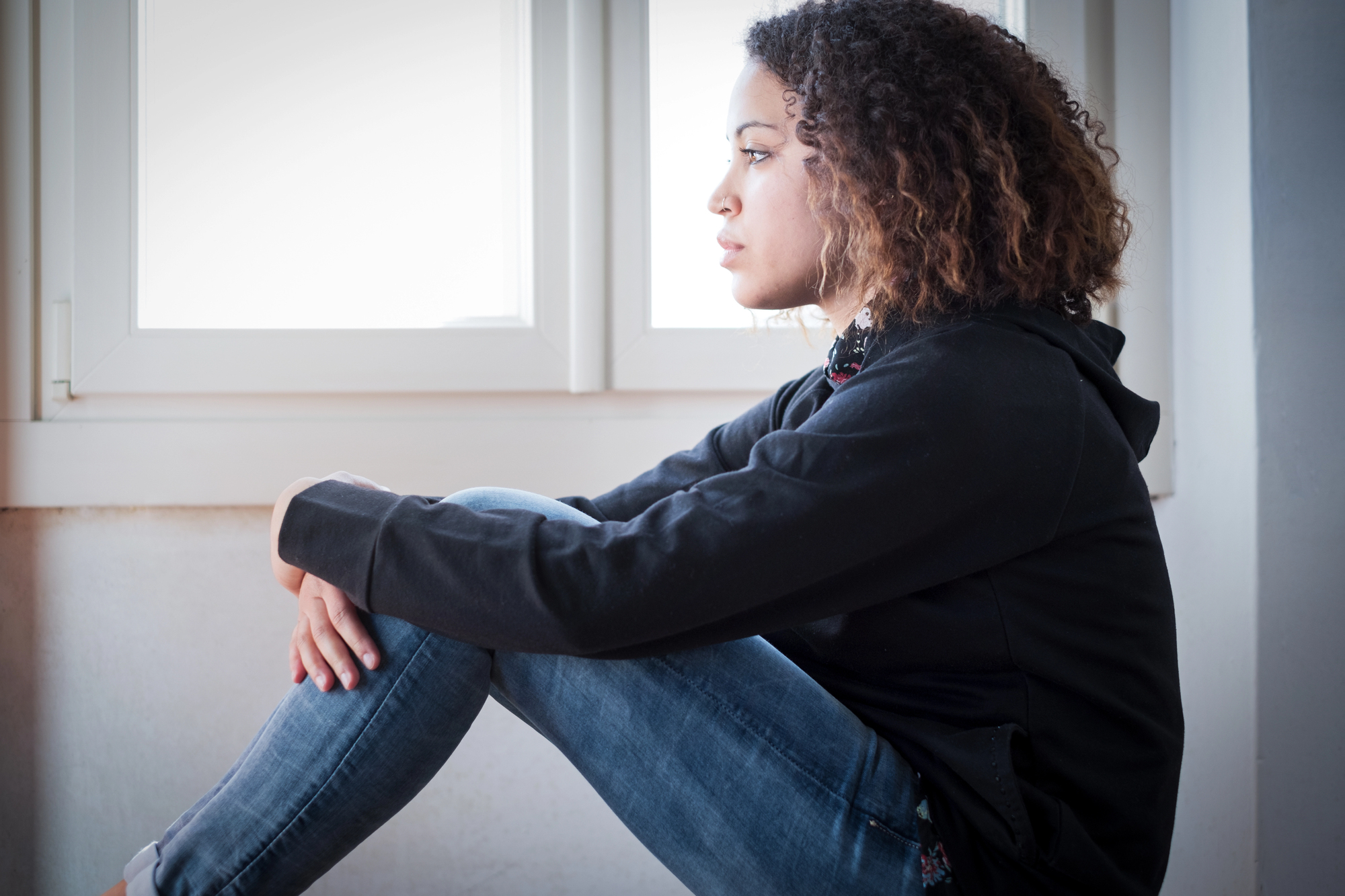 A person with curly hair, wearing a black hoodie and jeans, is sitting indoors by a window, looking outside thoughtfully. The natural light illuminates their profile, casting a calm atmosphere.