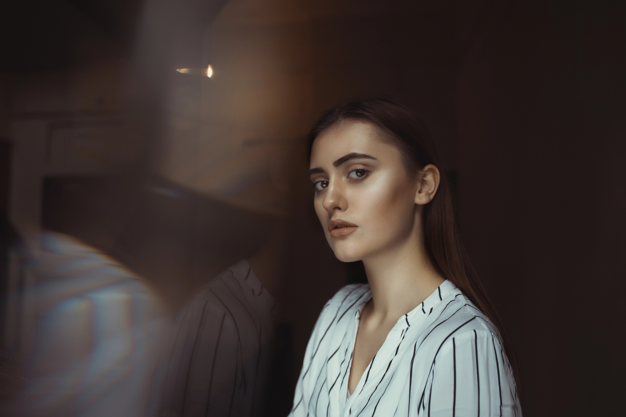 A woman with long brown hair and a serious expression gazes softly past the camera. She is wearing a white blouse with thin black stripes. The background is dimly lit, creating a moody atmosphere.