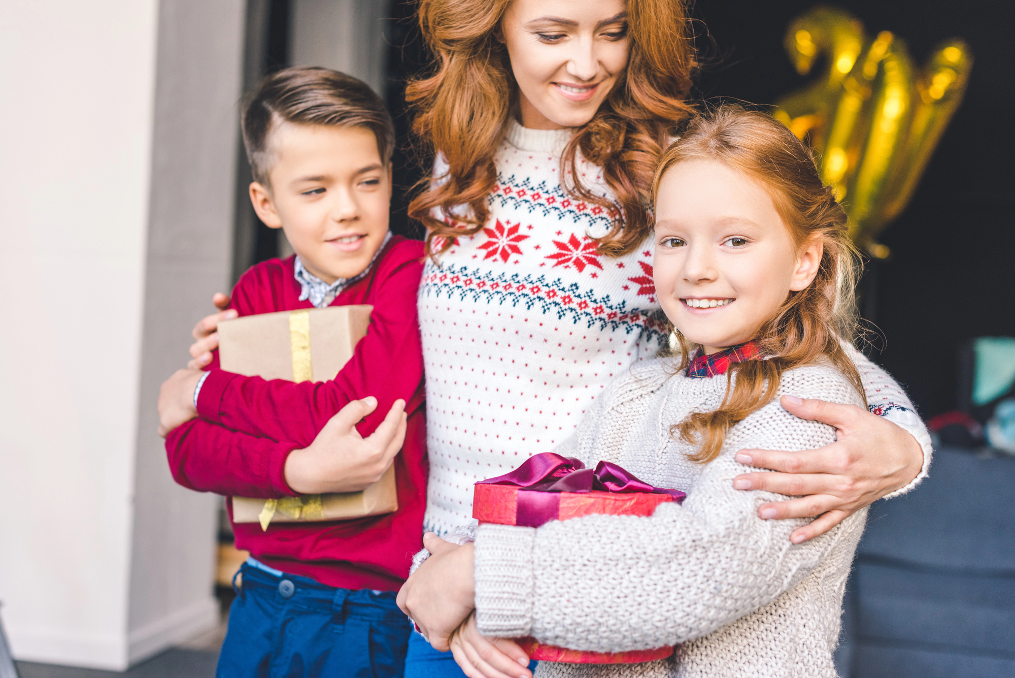 A woman and two children are indoors, smiling and holding wrapped gifts. The woman wears a festive sweater, the boy in a red sweater holds a gift with a gold ribbon, and the girl in a light sweater holds a pink present. Gold balloons are in the background.