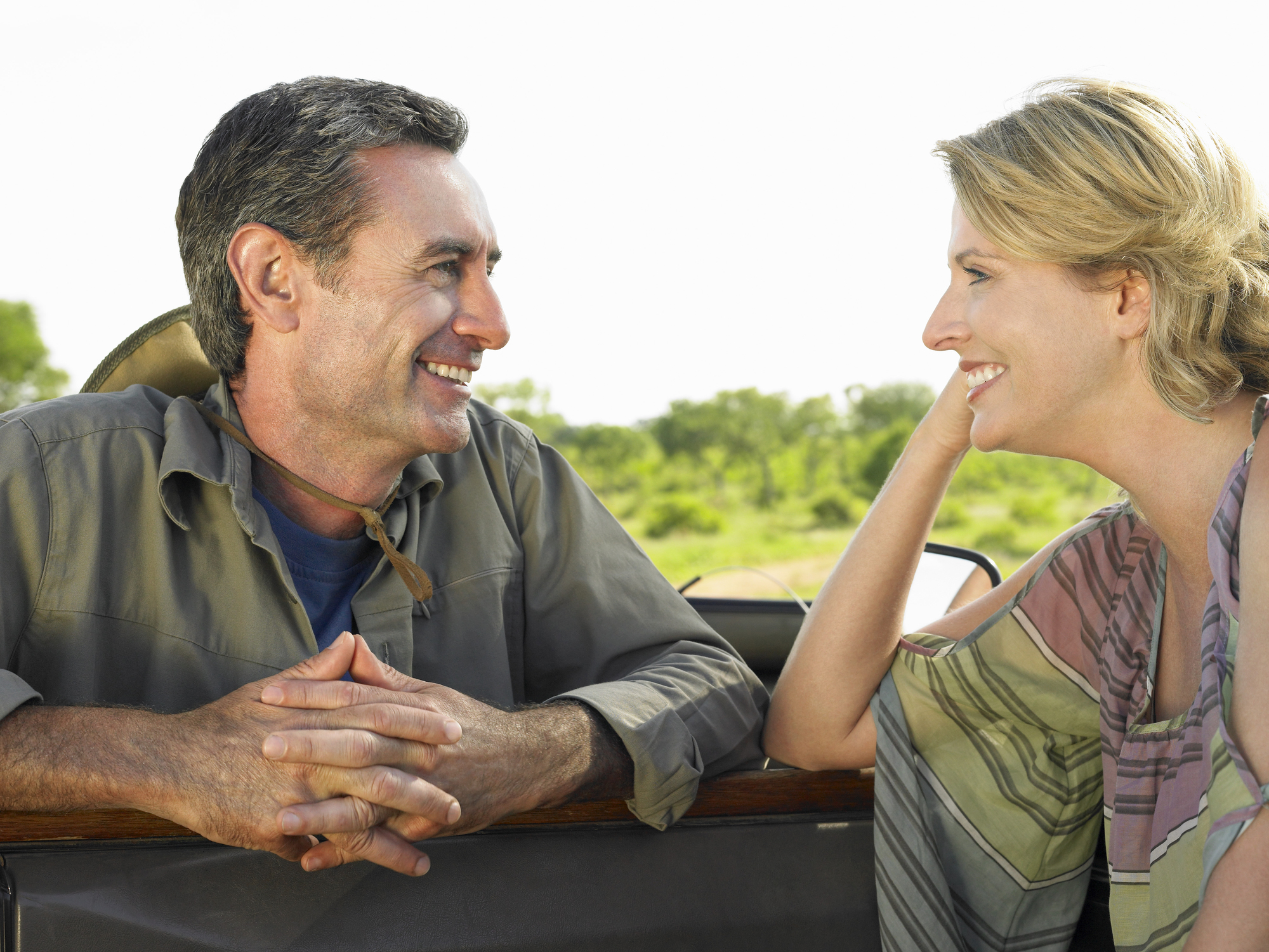 A man and woman smile warmly at each other while leaning on the side of a vehicle. The sunny outdoors and blurry greenery in the background suggest they are on a safari or nature excursion.