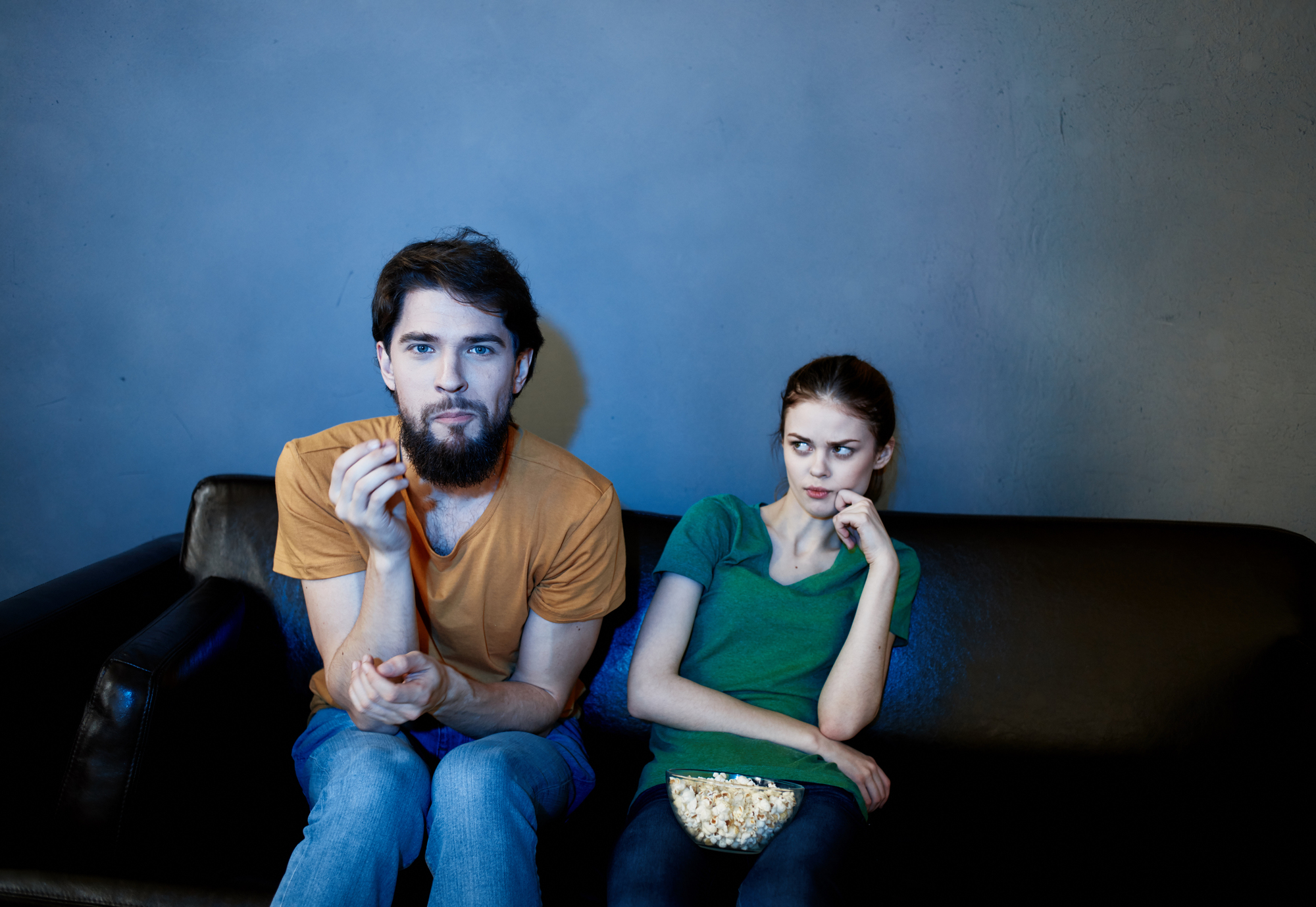 A man and a woman sitting on a couch watching TV. The man, in an orange shirt, is eating popcorn, while the woman, in a green shirt, looks at him skeptically. A bowl of popcorn is on her lap. The room is dimly lit.