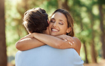 A woman with long brown hair smiles with her eyes closed, embracing a man outdoors in a sunlit forest. The background is blurred, with trees visible. The woman is wearing a sleeveless top, and the man is in a light blue sweater.