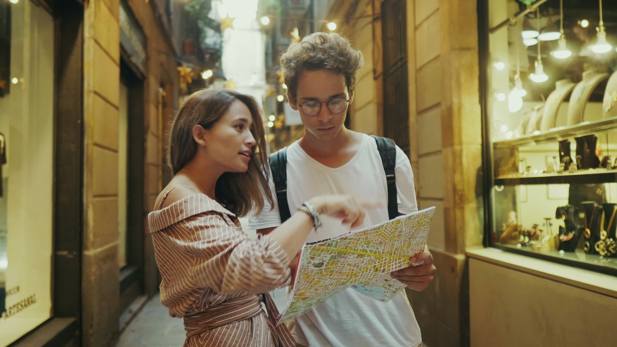 A woman and a man stand in a narrow alleyway between buildings. The woman, wearing a striped dress, points to a large map held by the man with glasses and a backpack. Shop windows line the walls, displaying various items. They appear engaged in conversation.