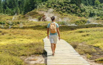 A person with a backpack walks on a wooden path surrounded by green vegetation and steam vents. Pine trees and a misty atmosphere create a serene, scenic backdrop.