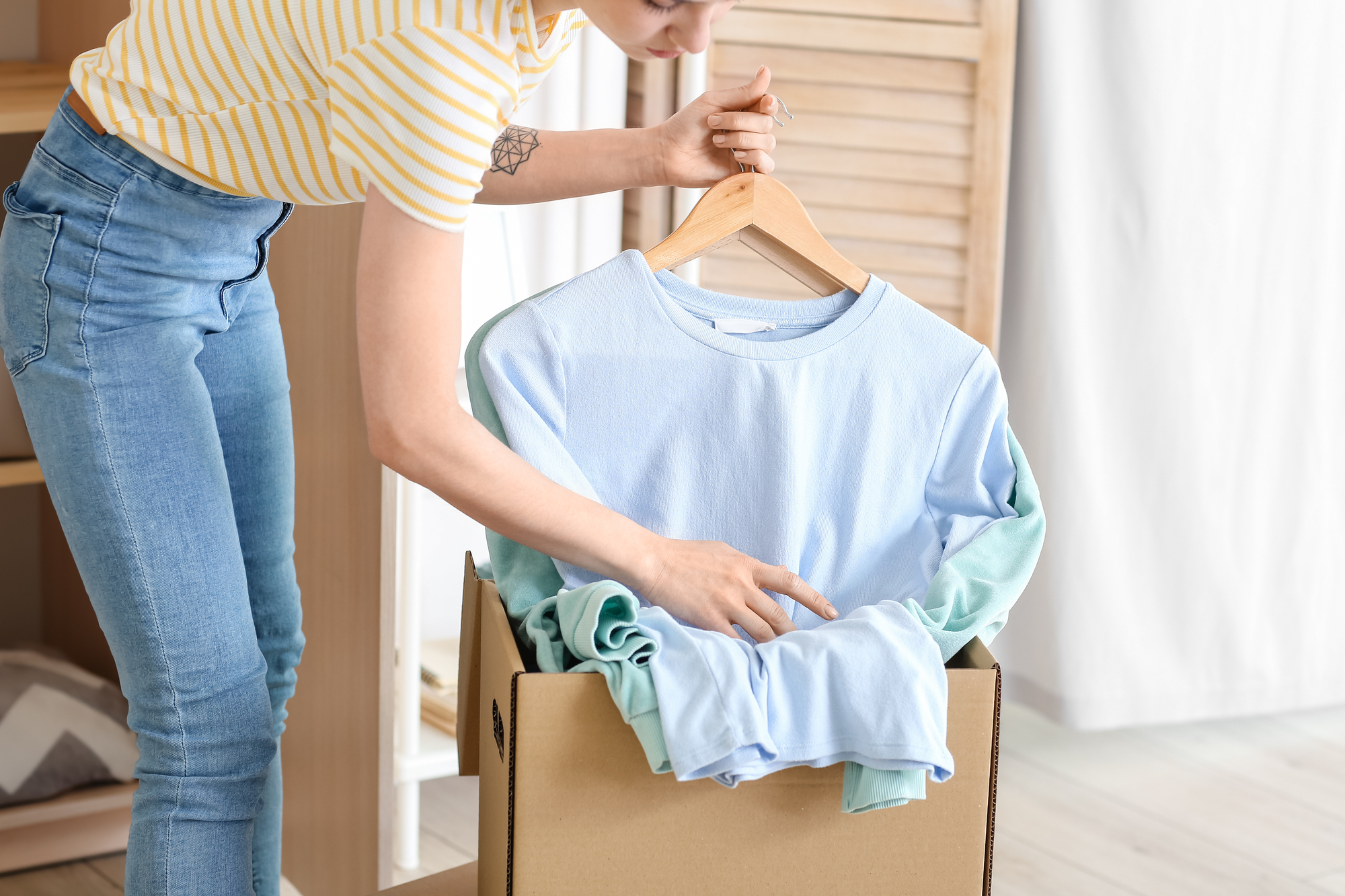 A person wearing a striped shirt and jeans places a pastel-colored long-sleeved shirt on a hanger. The shirt is partially inside a cardboard box, suggesting packing or unpacking. A closet with clothes is visible in the background.