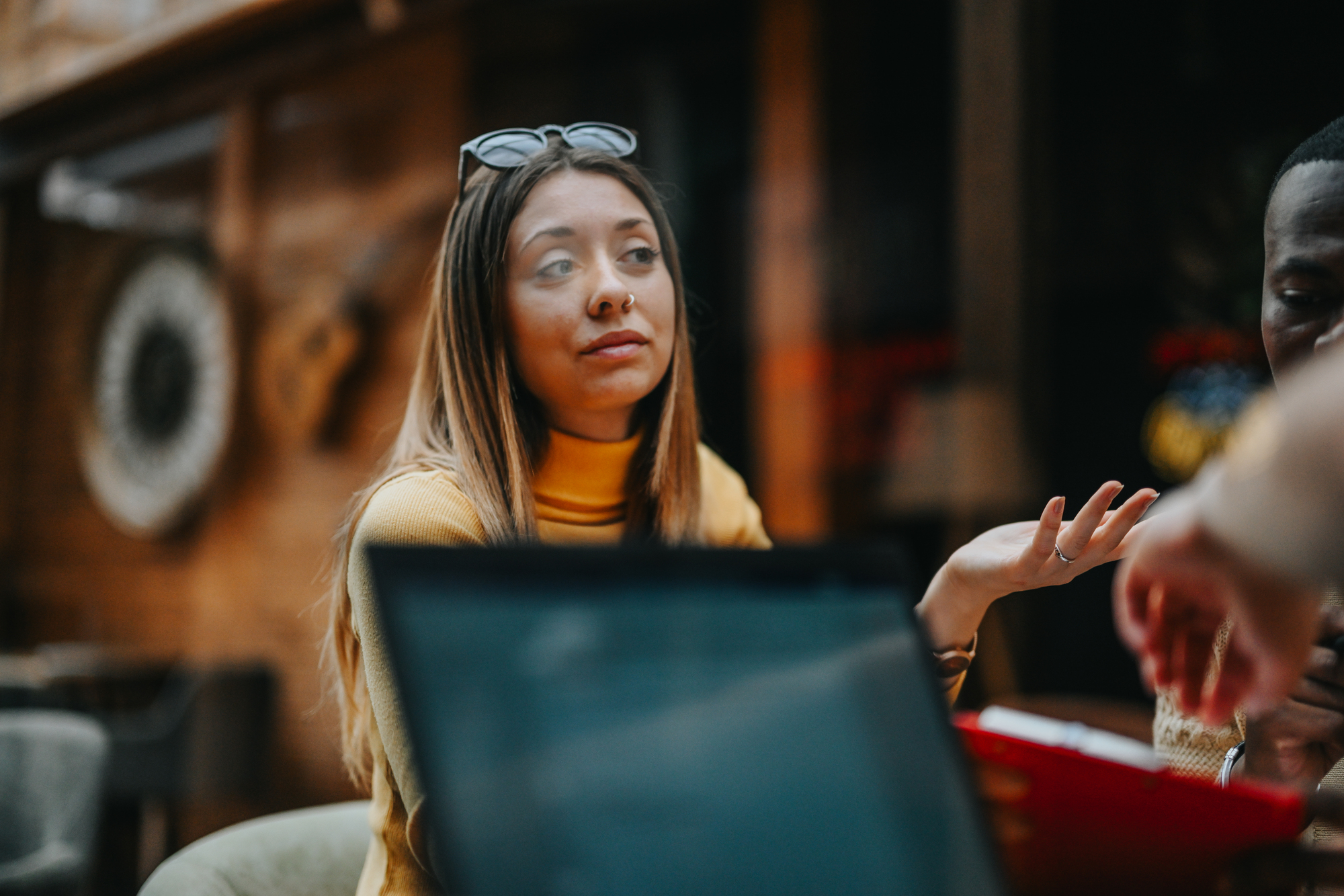 A woman with long hair and sunglasses on her head sits indoors, gesturing with one hand as she listens to someone off-camera. A laptop and another person are partially visible in the foreground. The setting has a cozy, wooden interior.