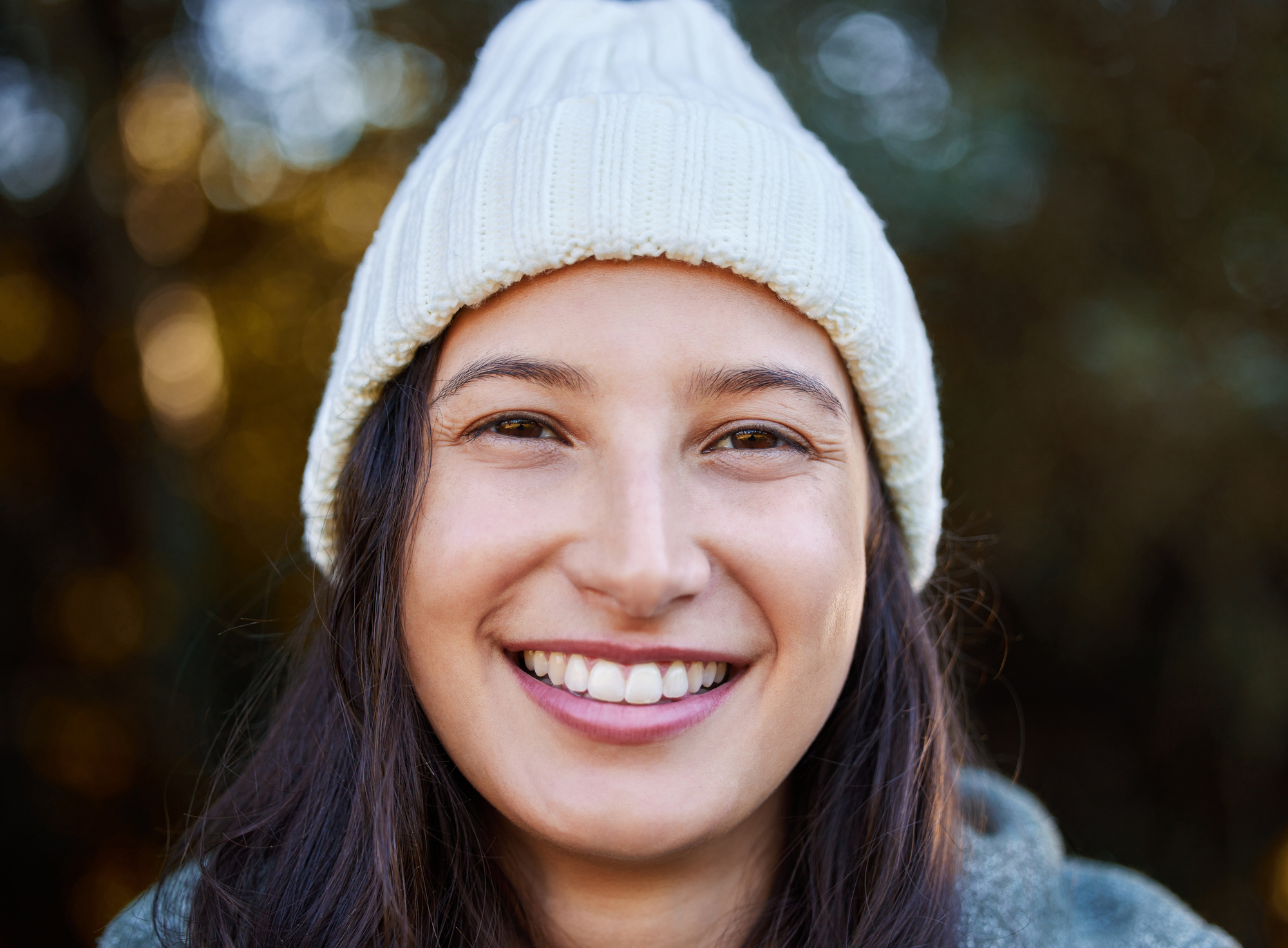 A person wearing a white knit beanie is smiling warmly at the camera. They have long dark hair and are standing outdoors, with a blurred natural background.