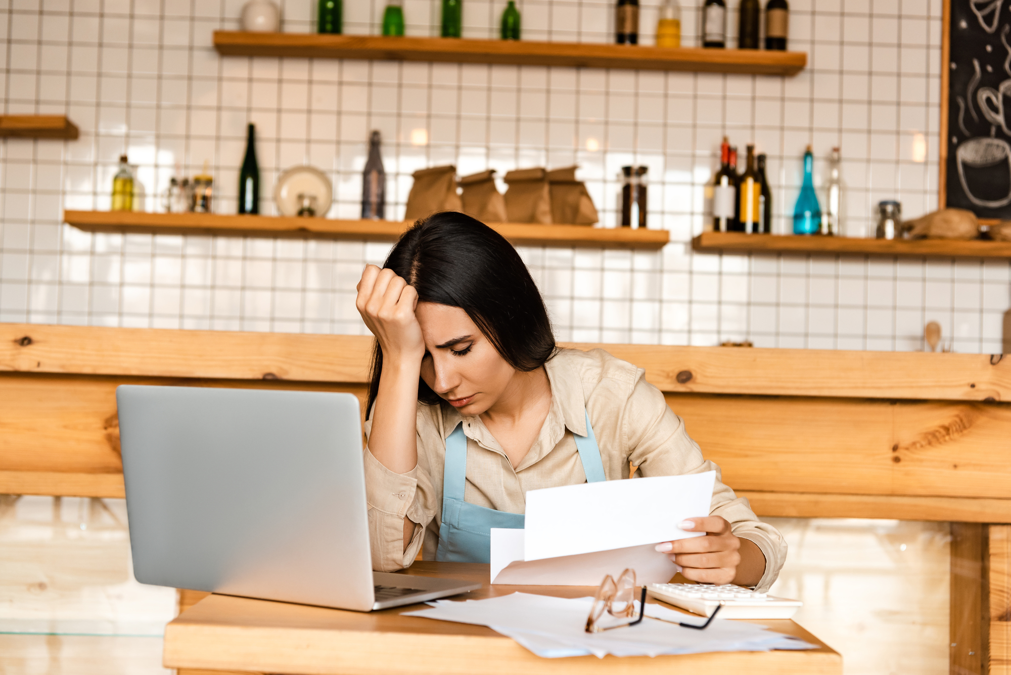 Woman sitting at a wooden table in a café, wearing an apron, looking stressed. She holds a piece of paper in one hand, with her other hand on her forehead. A laptop is open in front of her, surrounded by papers and glasses. Shelves with bottles in the background.