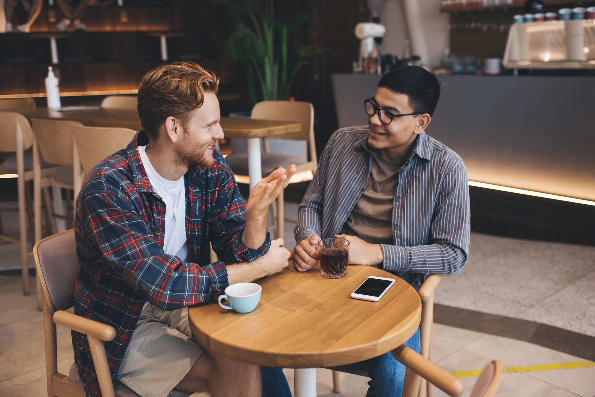 Two men sit at a wooden table in a coffee shop, engaged in conversation. One holds a phone on the table, and the other gestures with his hand. There are cups and glasses in front of them, and the background shows bar seating and a plant.