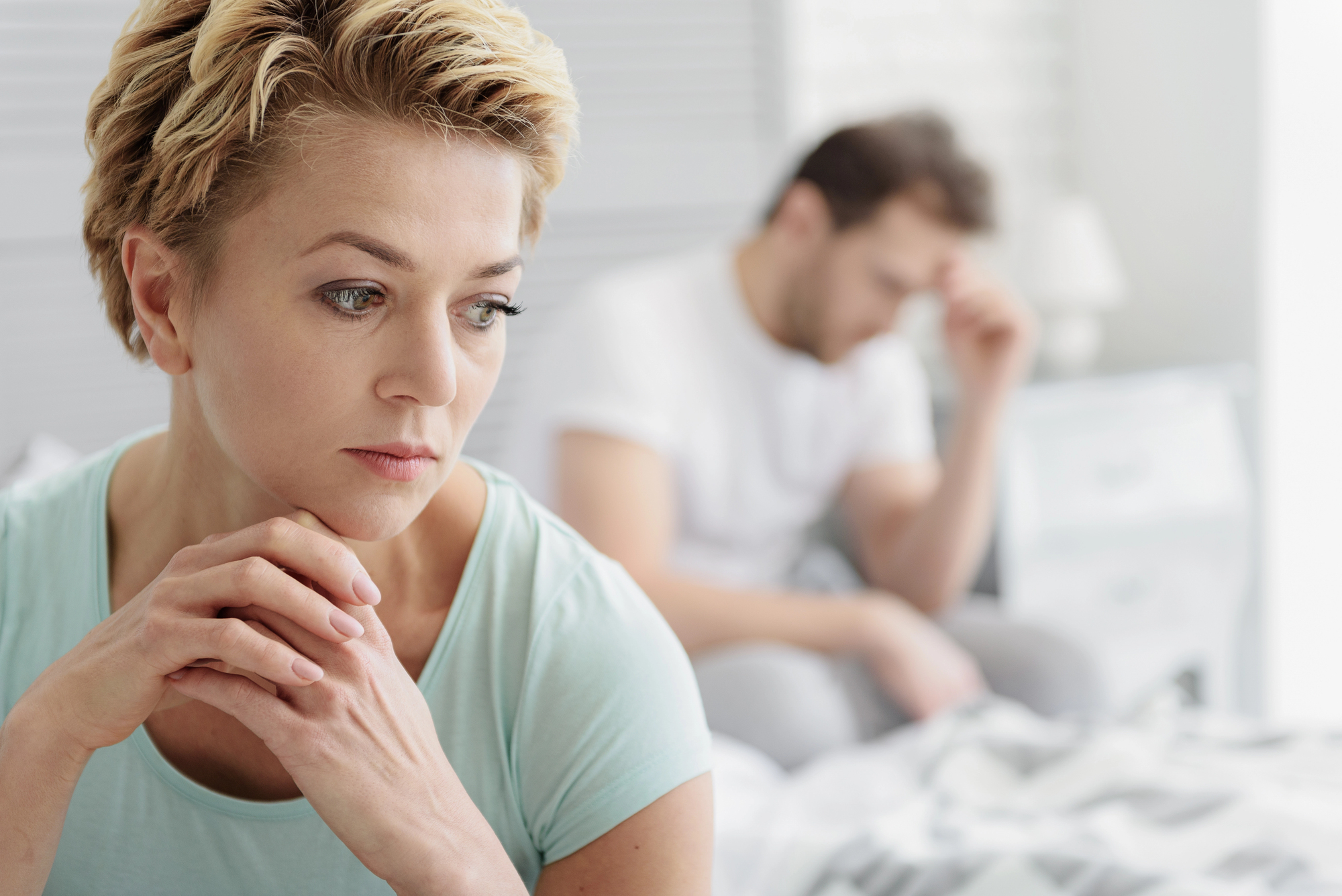 A woman with short blonde hair sits pensively on a bed, resting her chin on her hands. In the background, a man in a white shirt sits with his hand on his forehead, appearing distressed. The scene suggests tension or a serious conversation.