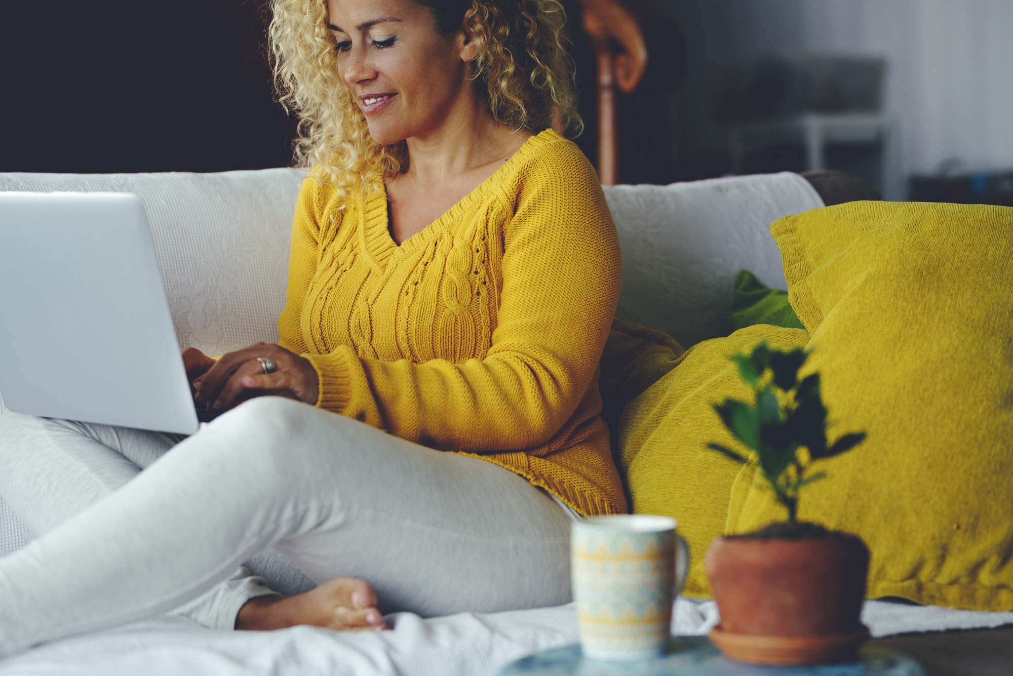 A woman with curly hair sits on a white sofa using a laptop. She is wearing a yellow sweater and white pants. In the foreground, there's a potted plant and a mug on a small table. Bright pillows are placed beside her.