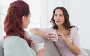 Two women sitting on a couch are having a conversation while holding white mugs. One has red hair in a bun, and the other has long brown hair. They are in a bright room with white curtains, dressed casually.