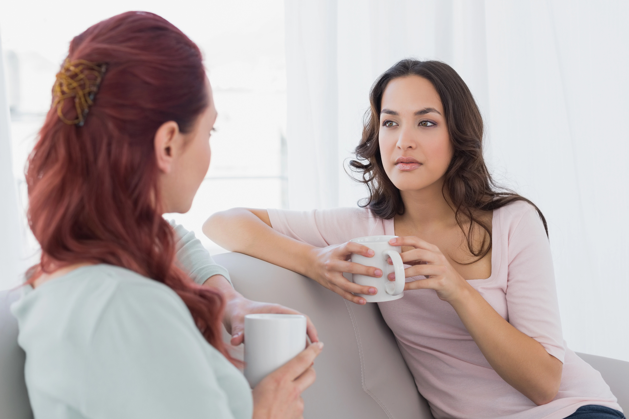 Two women sitting on a couch are having a conversation while holding white mugs. One has red hair in a bun, and the other has long brown hair. They are in a bright room with white curtains, dressed casually.