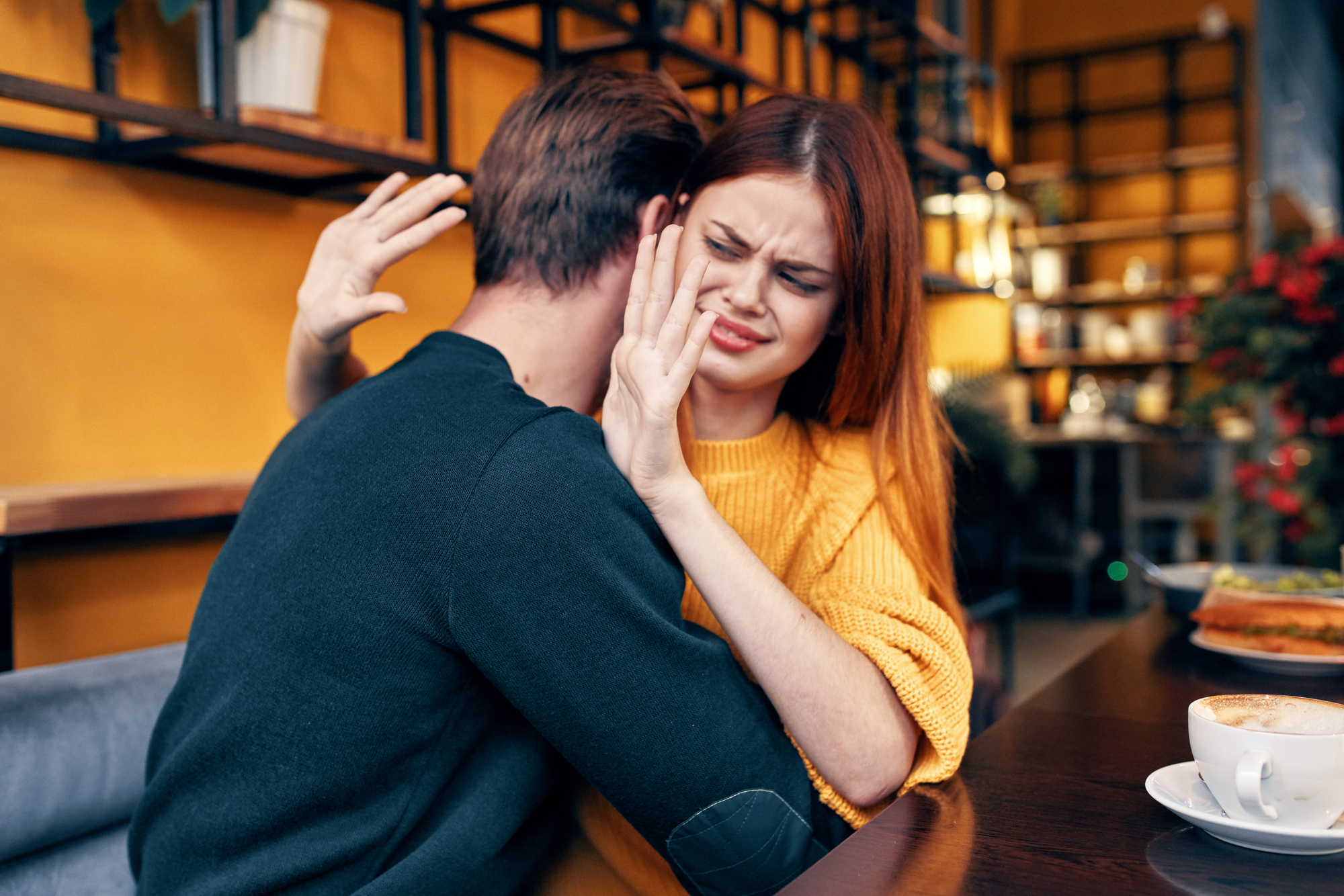 A woman with long red hair is sitting at a table in a cafe. She appears uncomfortable or upset as a man embraces her. Her hands are raised, and she is pressing her lips together. A cup and a plate are visible on the table.