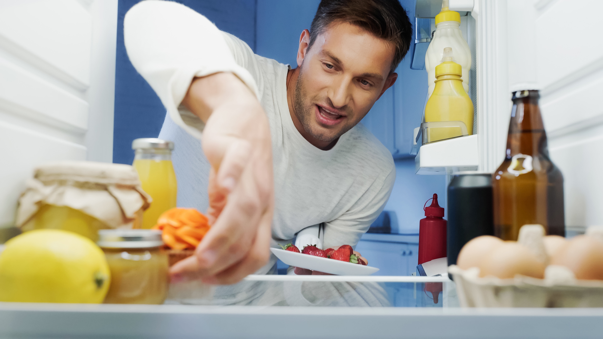 A man reaches into a refrigerator, smiling as he grabs a small container. Inside the fridge are eggs, a lemon, a jar of sauce, and various drinks. He's wearing a white shirt and looks content.