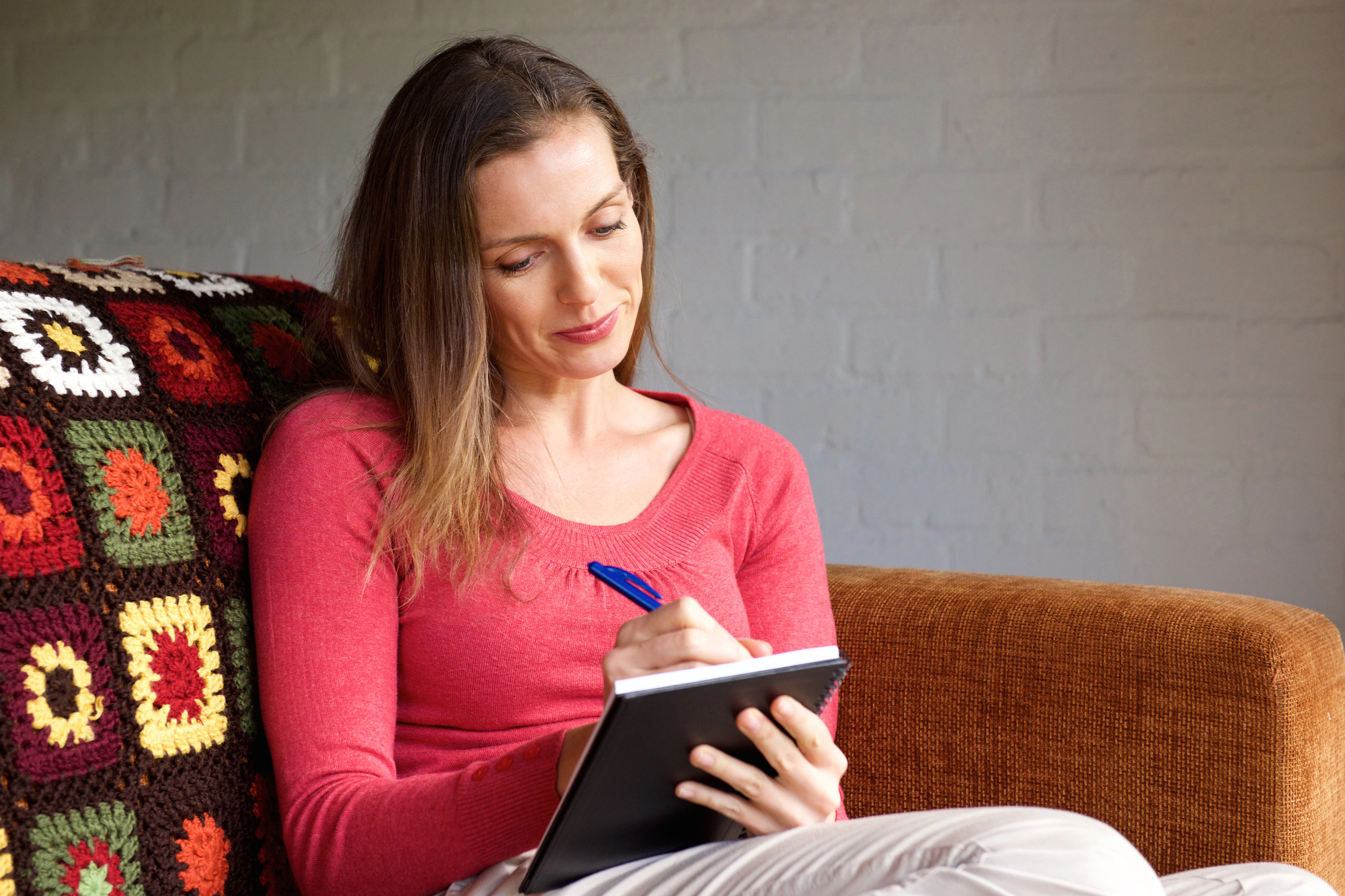 A woman sitting on a brown couch, wearing a pink sweater, is writing in a notebook with a blue pen. She appears relaxed and focused. The couch has a colorful crocheted blanket draped over its back. The background features a light gray wall.