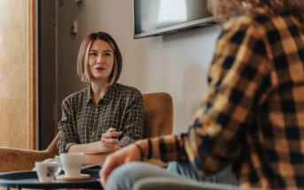 Two people are sitting in a cozy living room, engaged in conversation. One person holds a notebook and pen, while a coffee cup is on the table between them. They are both wearing checkered shirts, and there's a TV mounted on the wall.