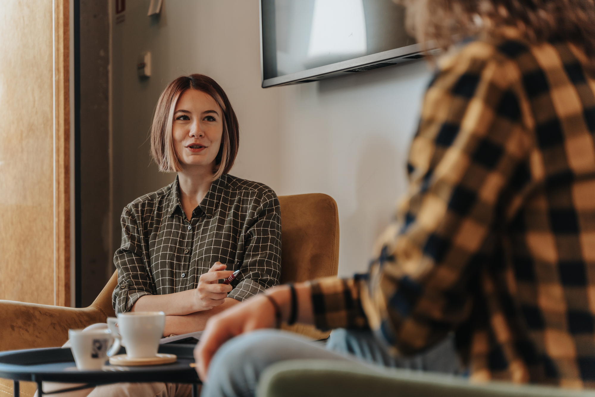 Two people are sitting in a cozy living room, engaged in conversation. One person holds a notebook and pen, while a coffee cup is on the table between them. They are both wearing checkered shirts, and there's a TV mounted on the wall.