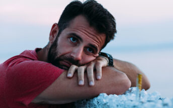 A man in a red shirt rests his head on his crossed arms, gazing contemplatively into the distance. He has dark hair and a beard, and a watch on his wrist. The background is blurred, suggesting a serene outdoor setting.