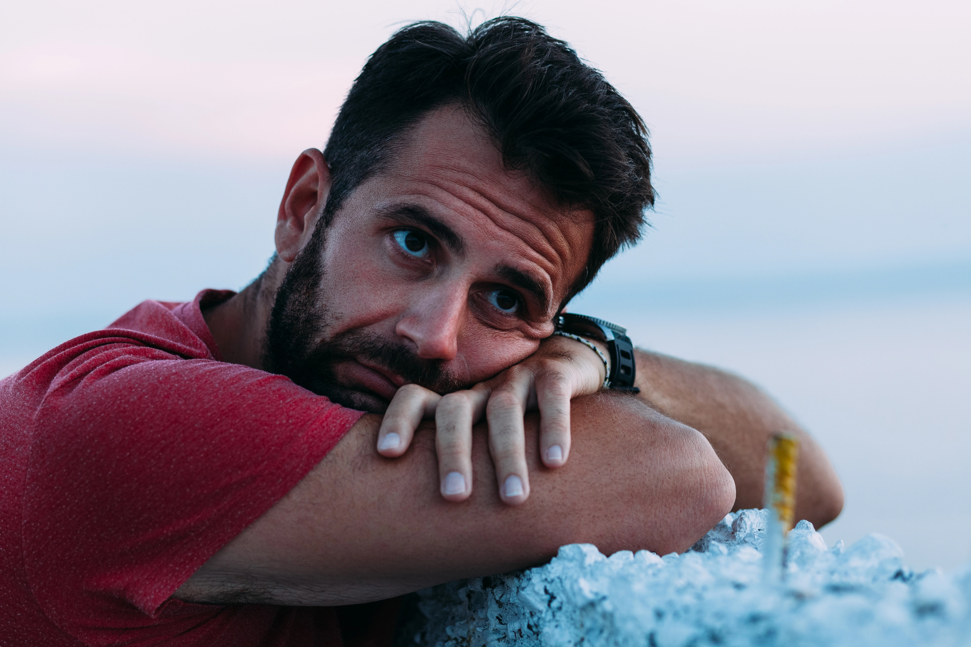 A man in a red shirt rests his head on his crossed arms, gazing contemplatively into the distance. He has dark hair and a beard, and a watch on his wrist. The background is blurred, suggesting a serene outdoor setting.